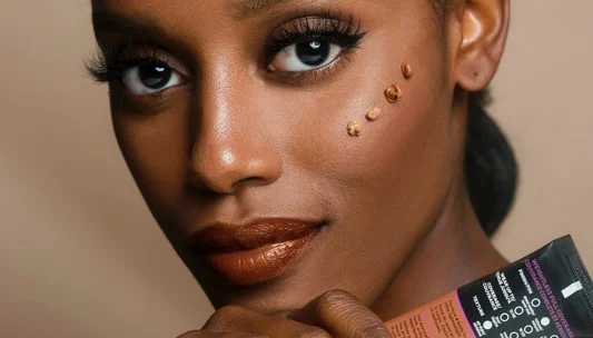 Close up of a dark skinned woman with makeup orange lipstick and small foundation dollops on her cheek while holding a cosmetic tube close to her face